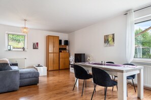 Ein modernes Wohnzimmer mit grauem Sofa, einem Esstisch mit Stühlen, einer Holzkommode und einem Fernseher.