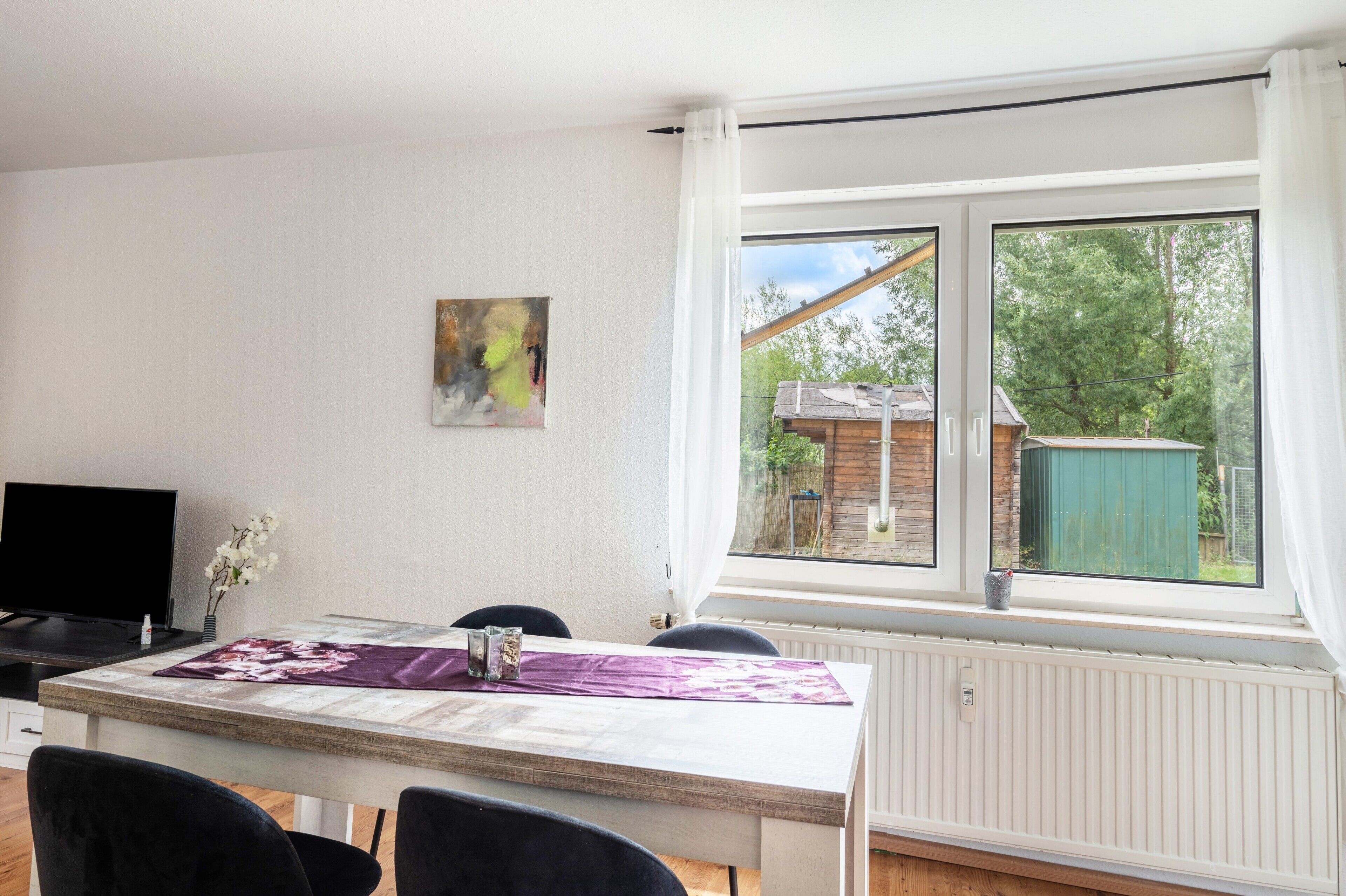 A dining area with a table, chairs, a television, and a window with a view of a wooden structure and greenery.