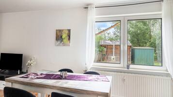 A dining area with a table, chairs, a television, and a window with a view of a wooden structure and greenery.