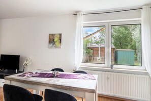 Een eethoek met een tafel, stoelen, een televisie en een raam met uitzicht op een houten constructie en groen.