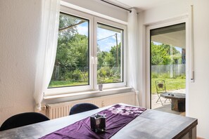 Een kamer met een tafel, stoelen, een raam met uitzicht op groen en een deur naar buiten.