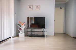 Un salon moderne avec un téléviseur à écran plat posé sur un meuble télé en verre, une bibliothèque remplie de livres et de magazines, et deux tableaux encadrés accrochés au mur.
