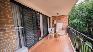 Uma varanda com piso de azulejos, uma cadeira de vime, uma mesa pequena com um vaso e um aparelho de ar condicionado branco.