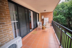 A balcony with a tiled floor, a wicker chair, a small table with a vase, and a white air conditioning unit. - Stylish Homebush Stay | Balcony, Parking, Sleeps 6 (Homebush West)