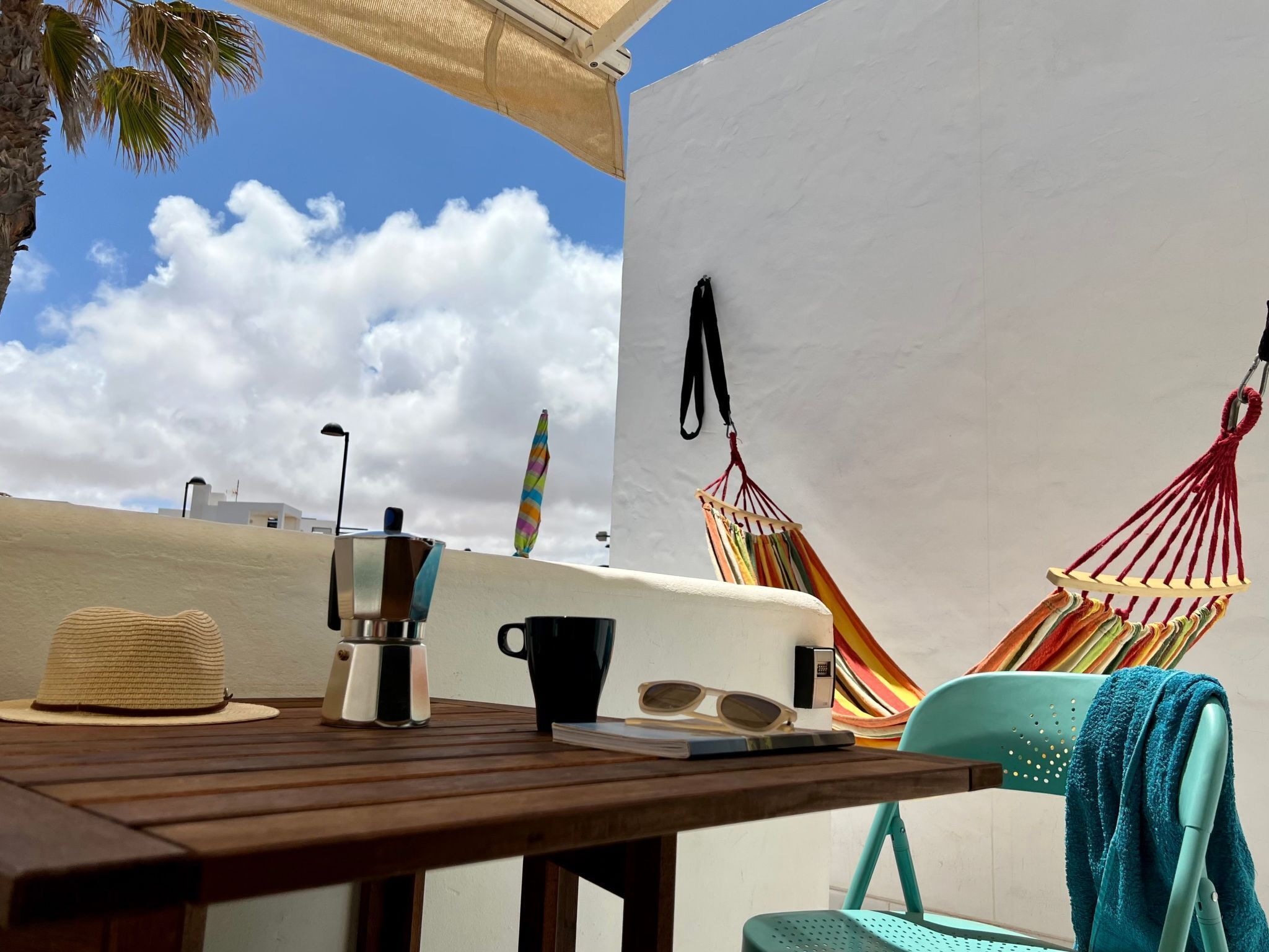 A wooden table with a coffee maker, a black mug, and a pair of sunglasses. There are two colorful hammocks and a teal chair with a towel.