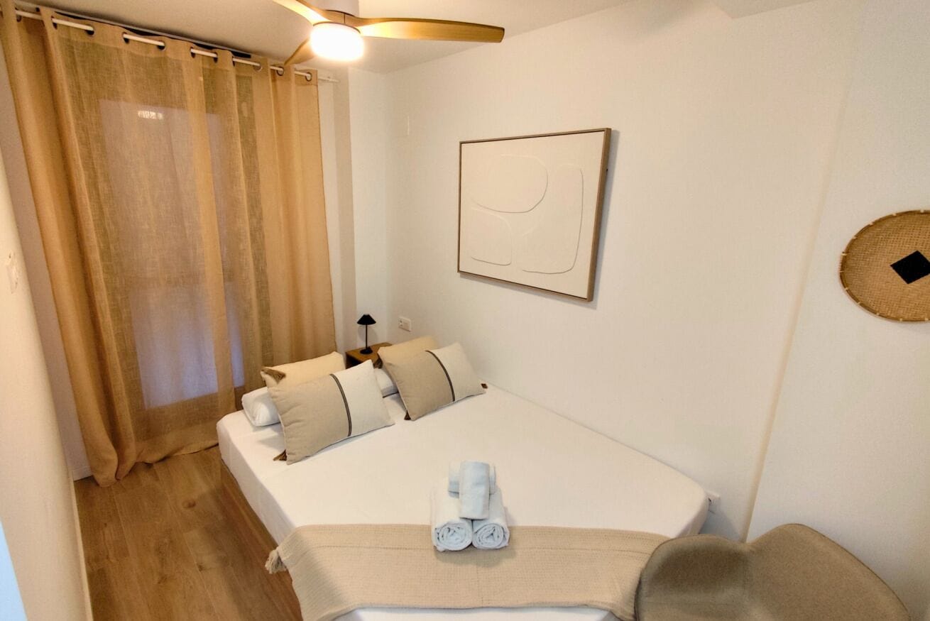 Een slaapkamer met een bed, nachtkastje, lamp, een aan de muur hangend kunstwerk en een gordijn.