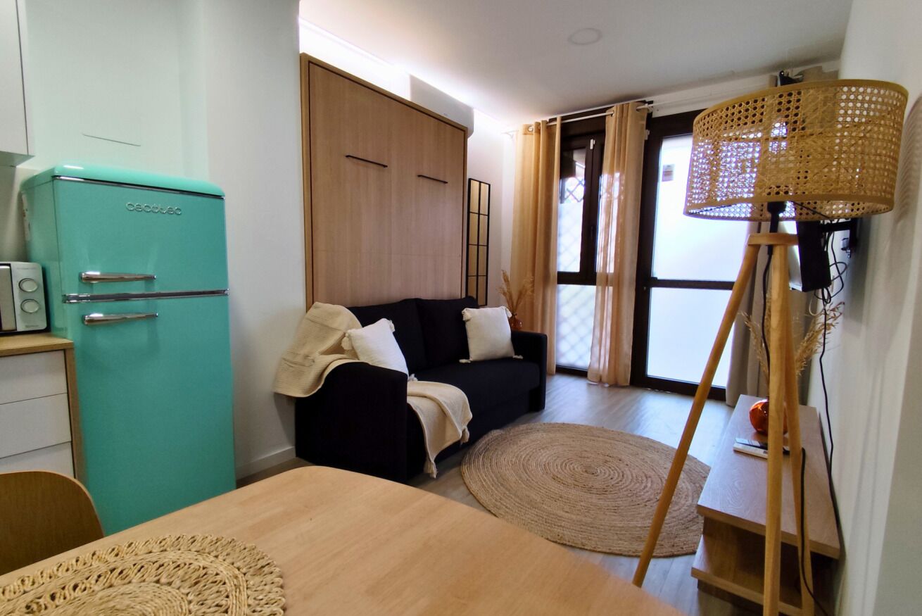 A compact room with a teal refrigerator, a black sofa, a wooden dining table, and a woven floor mat.