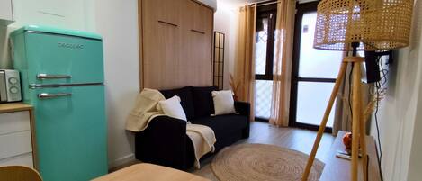 A compact room with a teal refrigerator, a black sofa, a wooden dining table, and a woven floor mat.