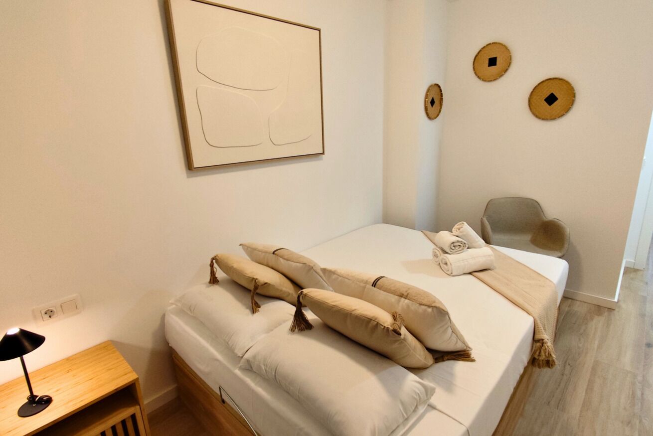 Una camera da letto ordinata con un letto, cuscini, una lampada da comodino, una sedia e un'opera d'arte appesa al muro.