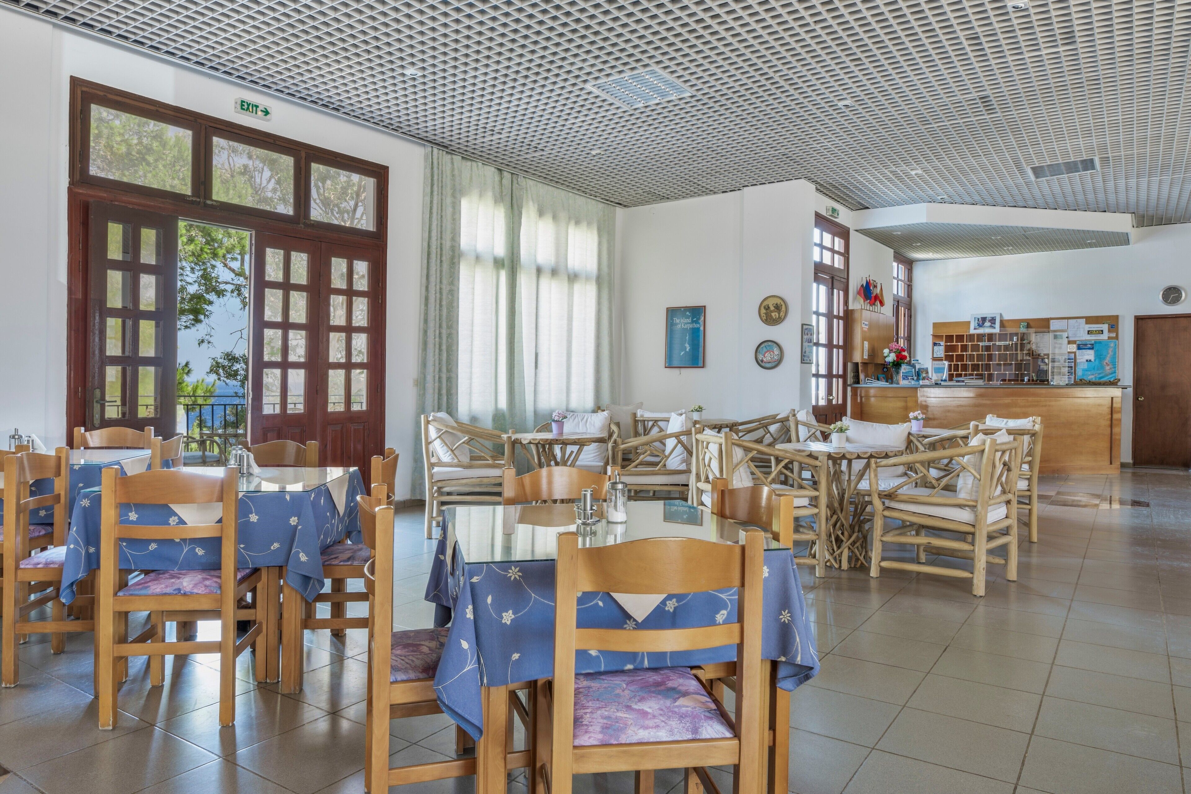 Een restaurantgedeelte in een hotel met meerdere tafels en stoelen, een vluchtwegbord en een receptiebalie.