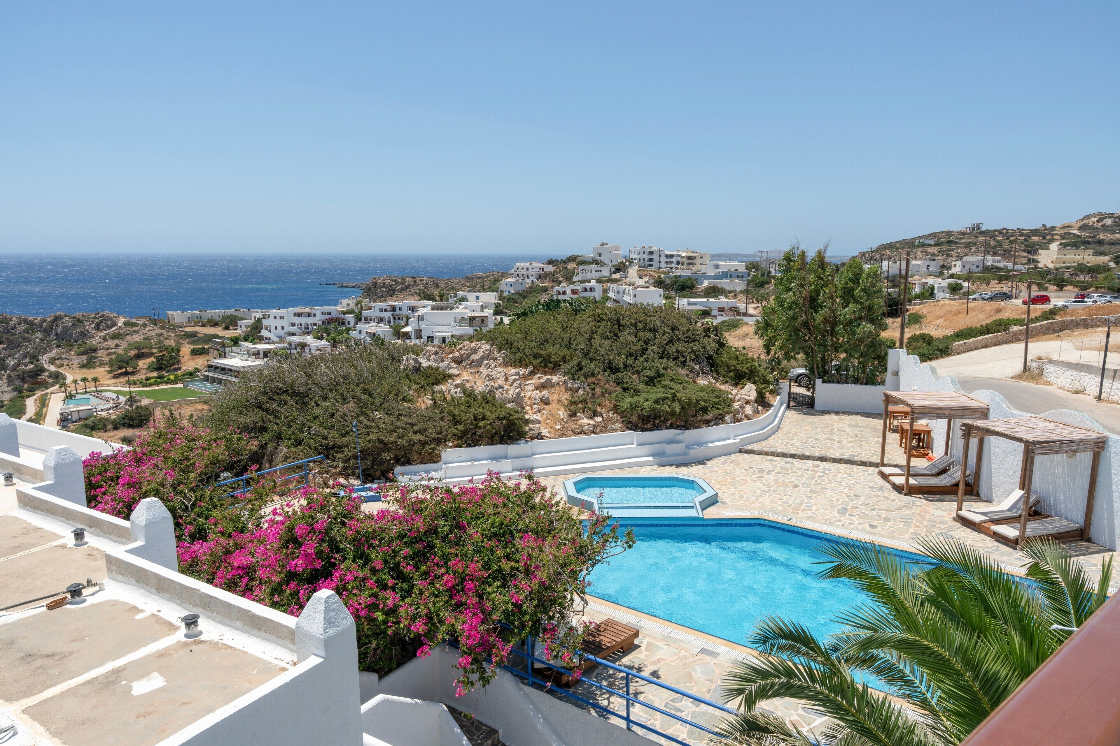 A pool with a view of the sea, surrounded by white buildings and greenery.