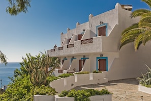 A coastal building with multiple balconies, a stone-paved courtyard, and lush greenery. - Standard Sea View Double 1 Room with Sea View and Shared Pool (Lakki)