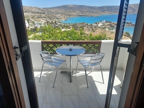 Un balcon avec une table et des chaises, donnant sur un paysage côtier avec des bâtiments et une étendue d’eau.
