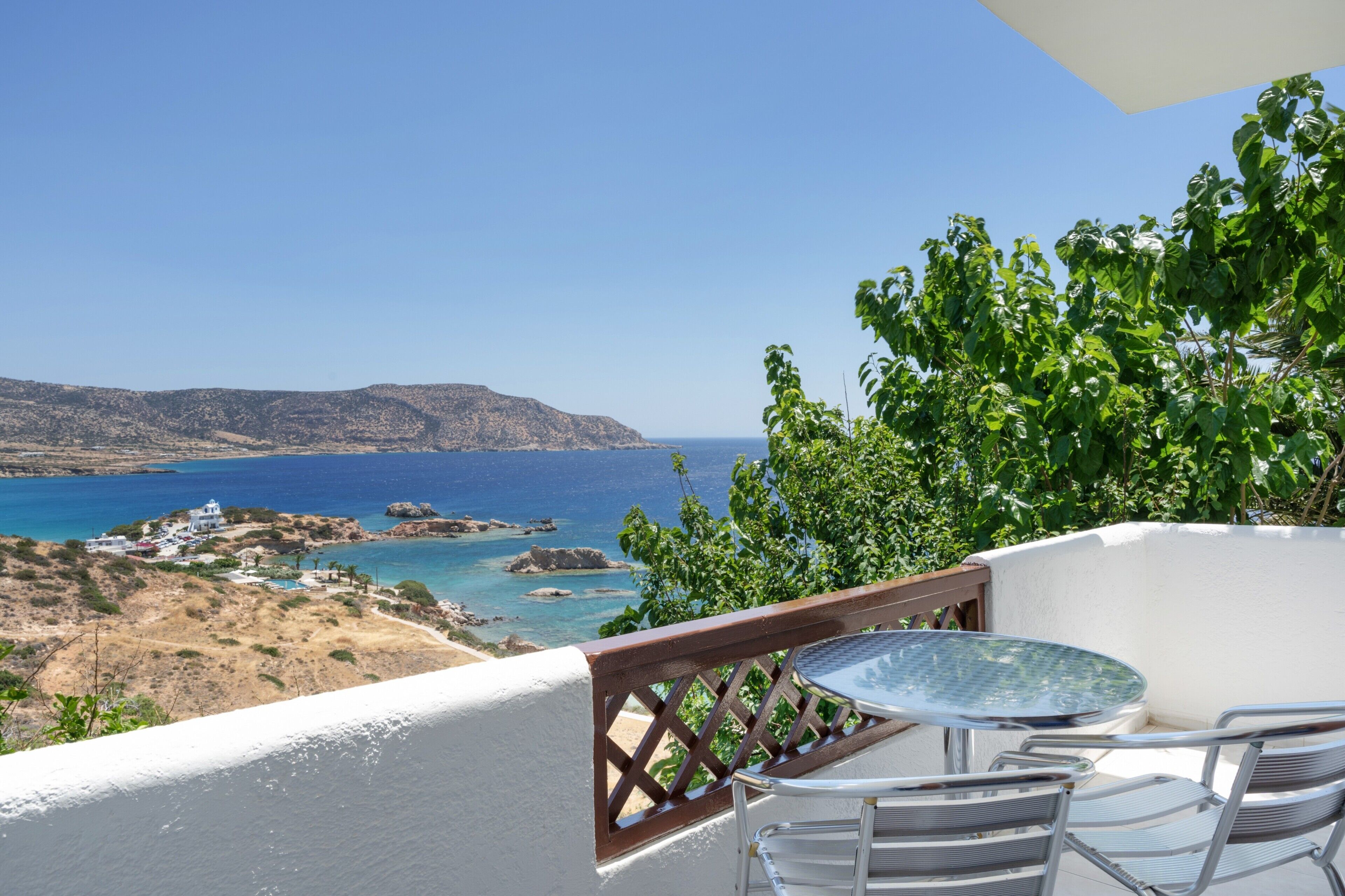 Un balcon avec une table et des chaises donnant sur un paysage côtier caractérisé par un terrain rocheux et une mer d’un bleu limpide.
