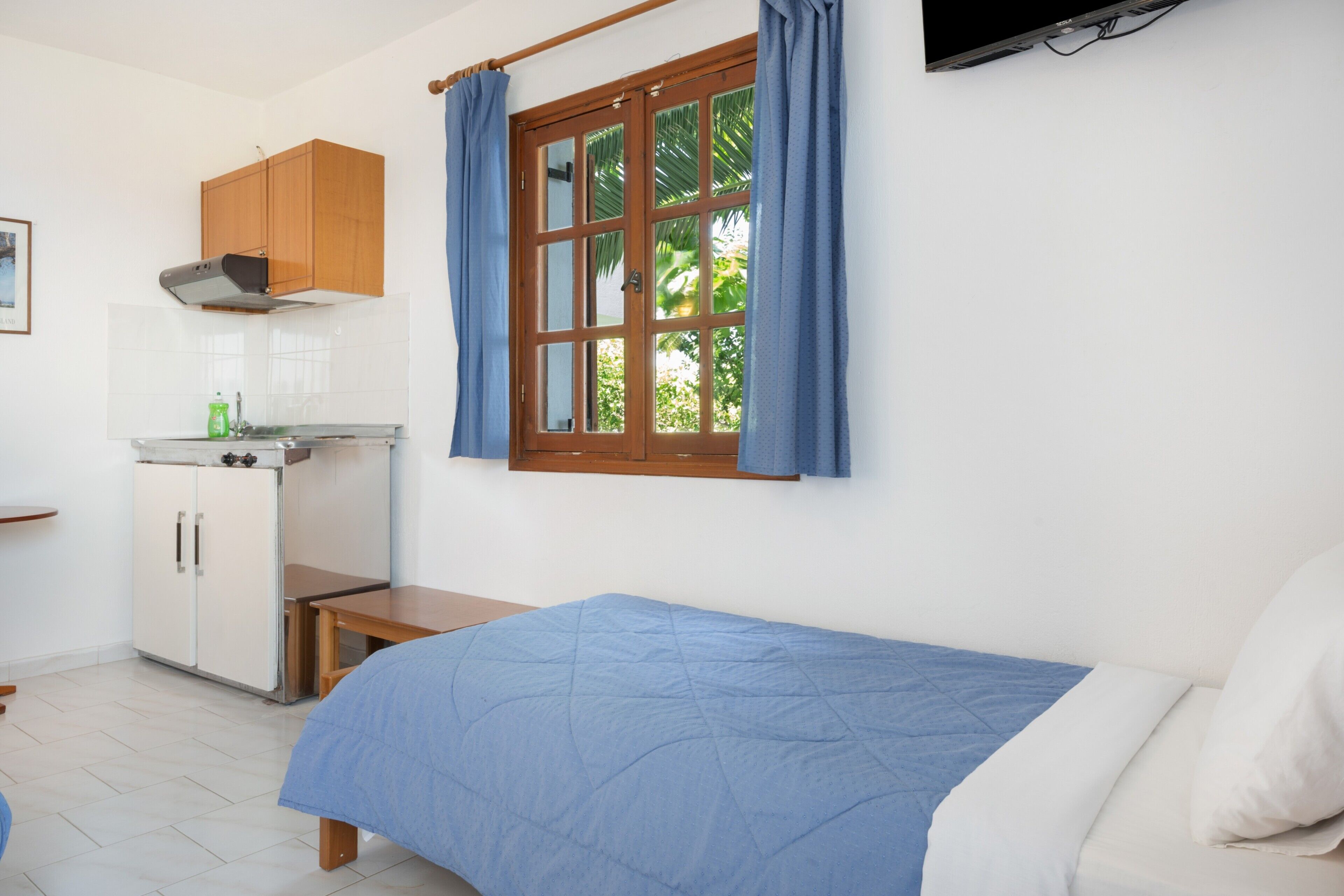 A bedroom with a bed, a window with curtains, a wooden cabinet, and a kitchenette.