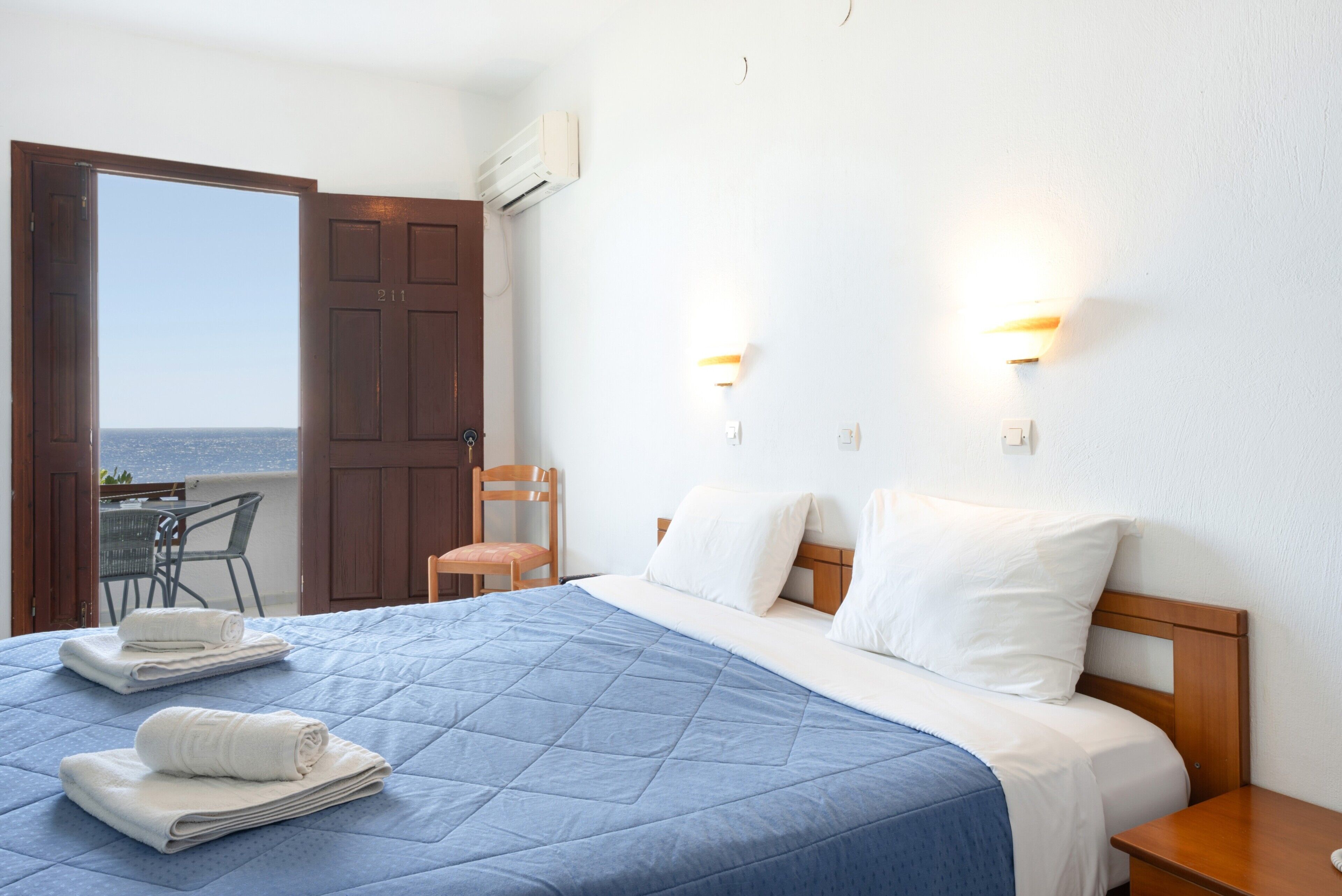 A hotel room with a bed, two nightstands, a chair, a table, and a view of the sea.