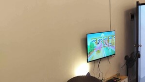 Quarto com televisão na parede, uma cadeira de vime, uma mesa redonda pequena e um tapete decorativo.