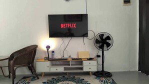 Quarto com televisão na parede exibindo 'NETFLIX', uma escrivaninha com computador, uma cadeira, um ventilador e um tapete decorativo.