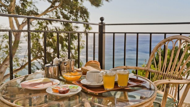 Une table en verre, sur laquelle sont disposés des aliments et des boissons pour le petit-déjeuner, se trouve sur un balcon donnant sur la mer.
