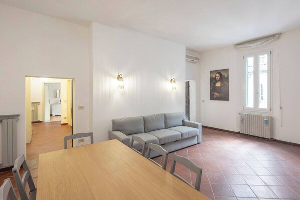A room with a dining table, chairs, a grey sofa, a painting on the wall, and a window.