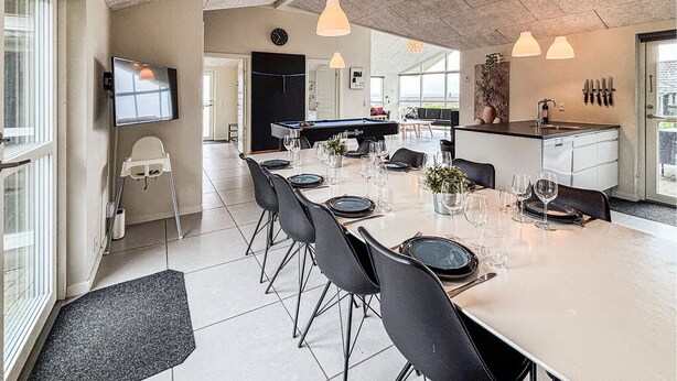Un espace repas moderne avec une longue table dressée pour six personnes, une table de billard et un coin cuisine équipé d’un évier et d’un plan de travail.