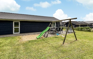 Ein Spielplatz mit Rutsche und Schaukeln, ein Gebäude mit dunkler Fassade und einem Ziegeldach, sowie eine Rasenfläche.