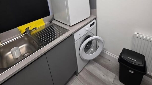 A compact kitchen with a refrigerator, washing machine, sink, and a black trash bin.