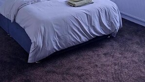 A bedroom with a bed, a nightstand, a window with blinds, and a radiator.