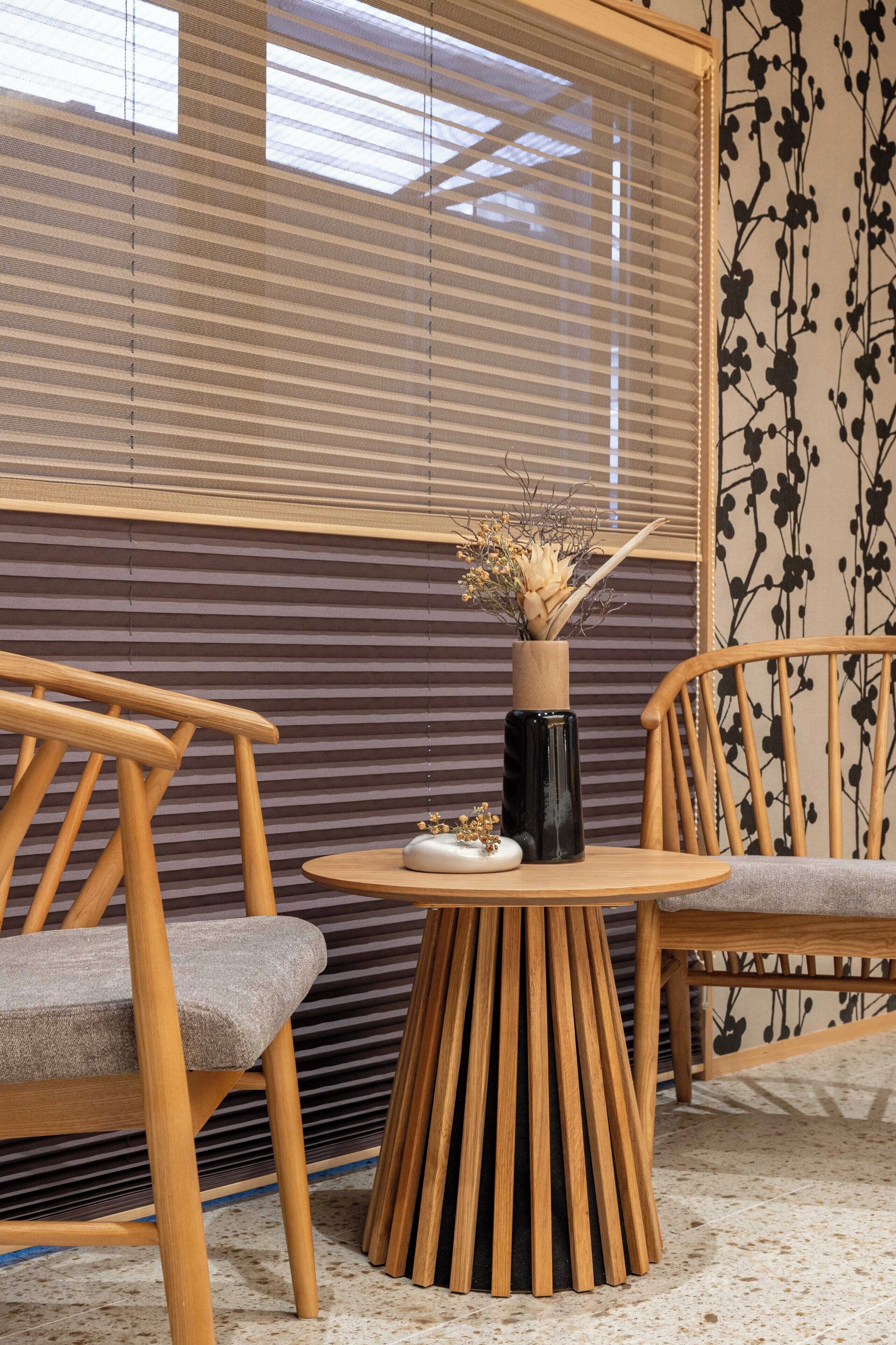 A room with a round wooden table, two wooden chairs, a vase with dried flowers, and a window with blinds.