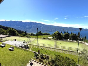 A tennis court with a seating area, surrounded by trees and mountains in the background. - Marcus 706 Apartment, Residence with Pool and Lake View (Tignale)