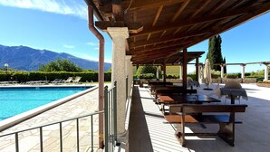 Un espace aménagé au bord de la piscine, doté d’un coin salon couvert, d’un sol en pierre et offrant une vue sur les montagnes en arrière-plan.