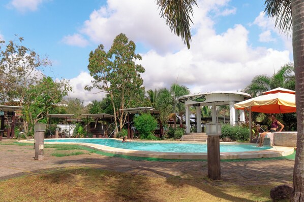 Una piscina con un pabellón, palmeras y una zona techada con una sombrilla.