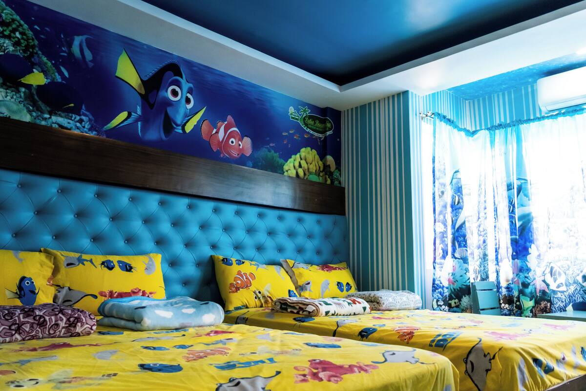 Habitación de hotel con un mural grande de vida marina, un cabecero capitonado y dos camas con ropa de cama colorida.