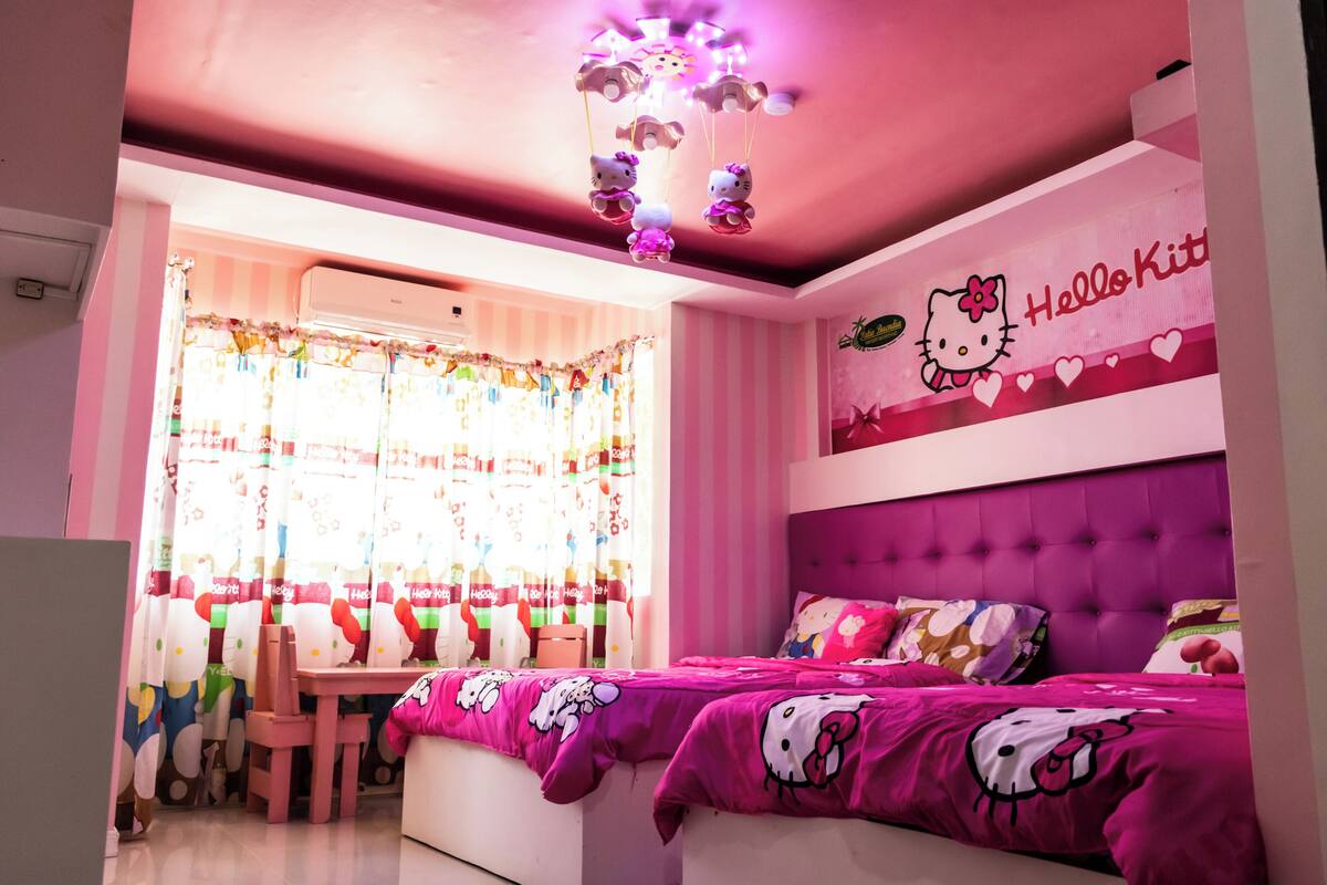 Una habitación con temática rosa, una cama, una ventana con cortinas y un letrero de Hello Kitty en la pared.