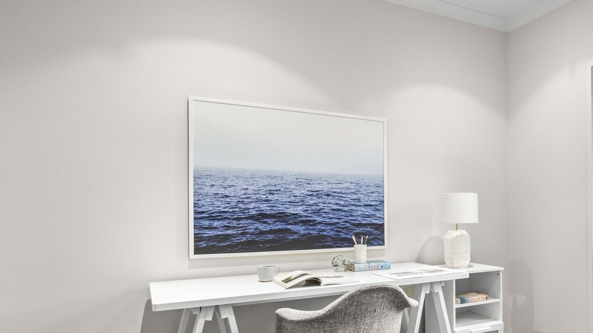 A white desk with a framed picture of the ocean, a lamp, and a chair.