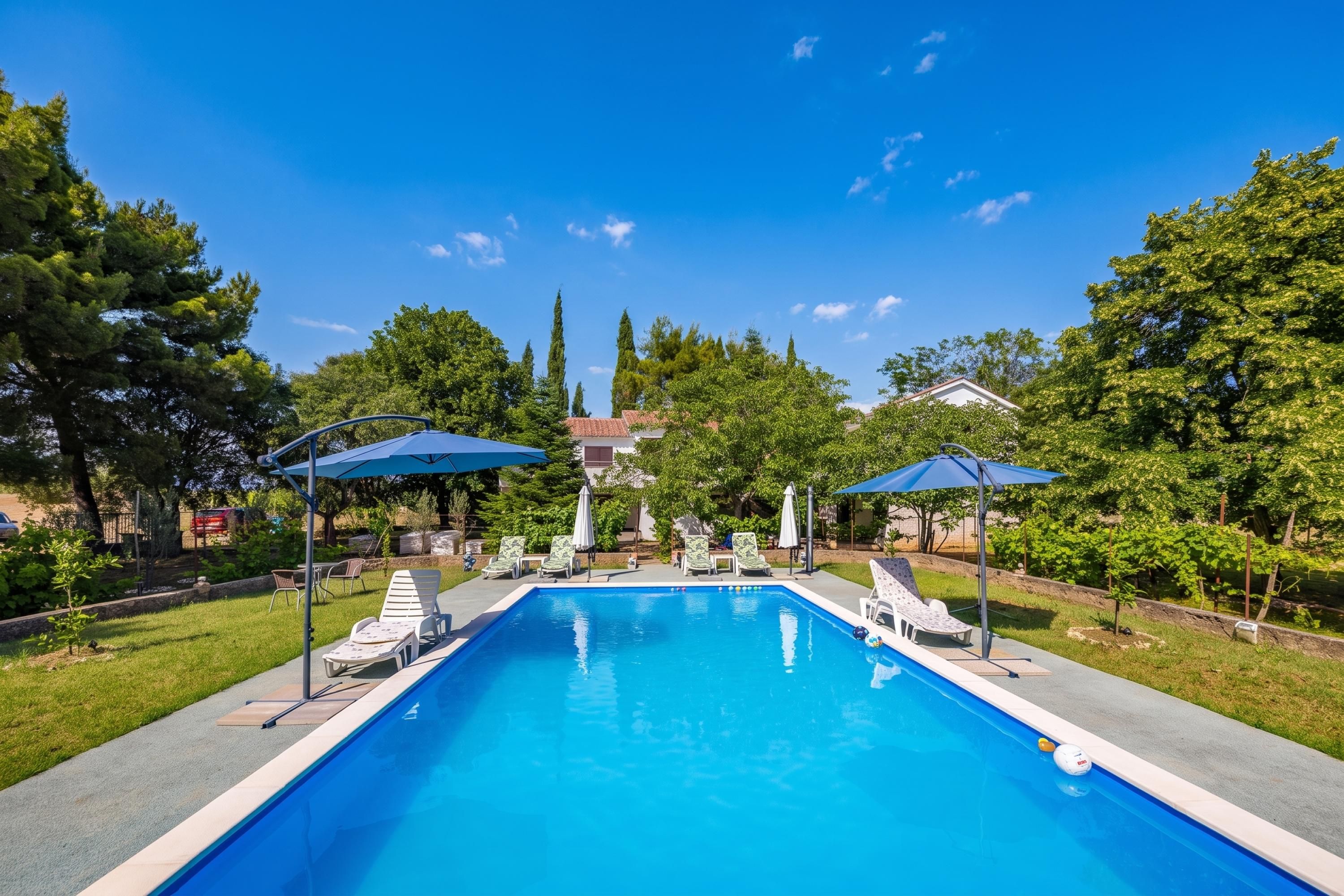Une piscine rectangulaire, dotée de chaises longues et de parasols, entourée d’arbres et avec un bâtiment visible au loin.