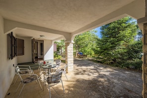 A covered patio with a table and chairs, a stone pillar, and a view of a tree-lined path. - Pool & Garden House Jaga (Polaca)