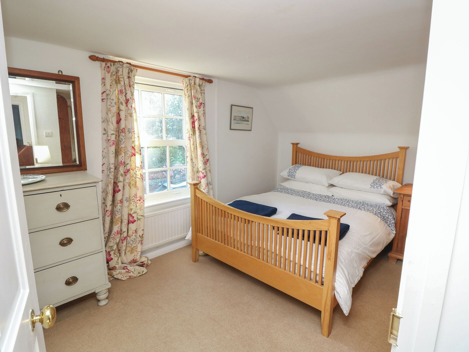 Une chambre à coucher avec un lit en bois, une commode à tiroirs, une fenêtre ornée de rideaux à motifs floraux et un miroir accroché au mur.