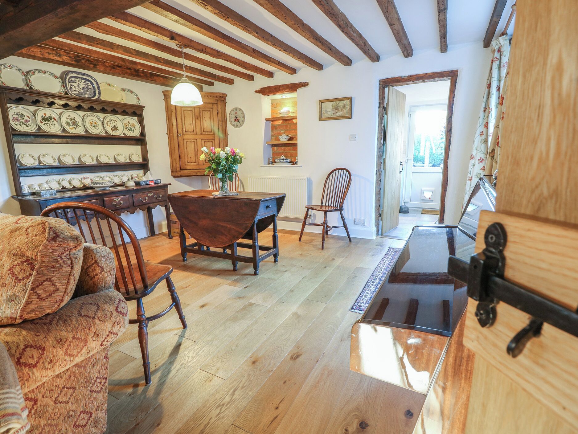 A rustic kitchen with wooden beams, a dining table, chairs, and a variety of dishes displayed on shelves.
