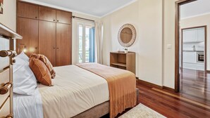A bedroom with a bed, wooden wardrobe, a round mirror, and a view of the kitchen. - Our Nest - Where Light and Tranquility Meet (Caniço)