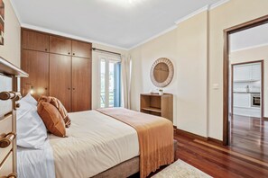 A bedroom with a bed, wooden wardrobe, a round mirror, and a view of the kitchen. - Our Nest - Where Light and Tranquility Meet (Caniço)