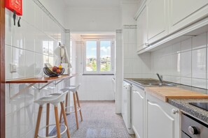 A narrow kitchen with white cabinets, a wooden countertop, and a window with a view of greenery. - Our Nest - Where Light and Tranquility Meet (Caniço)