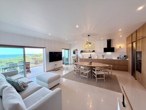 A modern living room with a large flat-screen TV, a dining area with a wooden table and chairs, and a kitchen with a built-in oven and refrigerator.