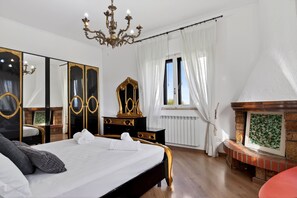 Una camera da letto con un letto, una cassettiera, uno specchio, un lampadario, un camino e una finestra con tende.