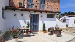 Un patio con una mesa redonda y sillas, plantas en macetas, y un edificio con fachada de ladrillo y puerta azul.
