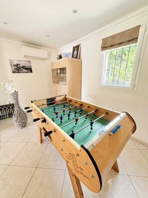 Une table de baby-foot dans une pièce dont le sol est carrelé, avec un meuble dont les portes sont en verre et une fenêtre donnant sur un paysage verdoyant.