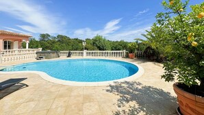 Una piscina de borde curvo, rodeada por una terraza con baldosas, una cerca blanca y una casa de fachada rosa al fondo.
