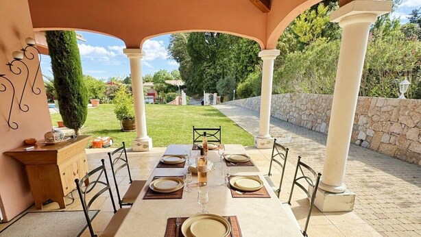 Un patio exterior con una mesa lista para comer, rodeado de sillas, bajo una zona cubierta con columnas.