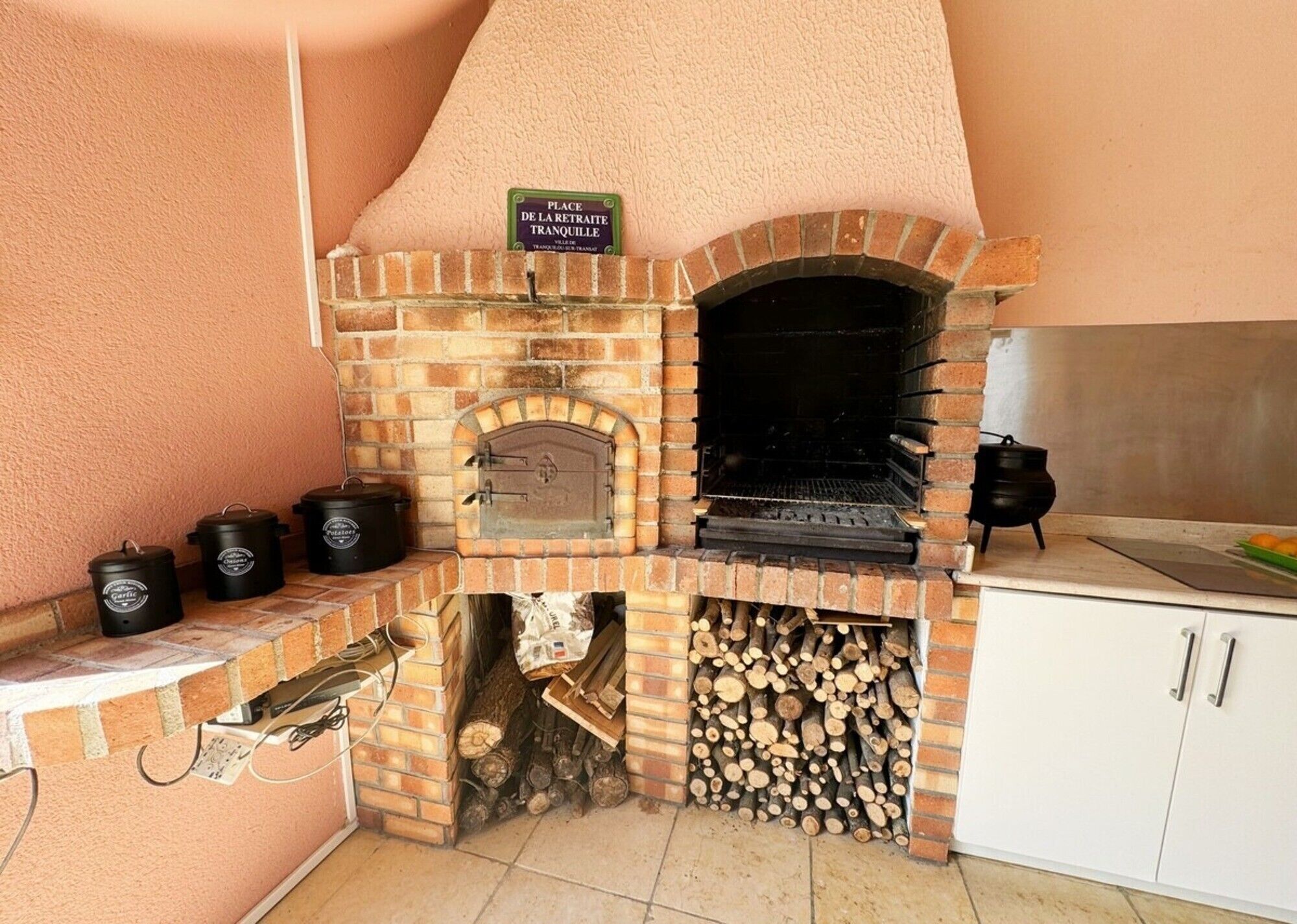 A brick-built outdoor oven with a sign above it, a stack of firewood, and a white cabinet to the right.
