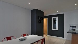 A modern dining area with a white table and red chairs, a dark accent wall, and a kitchen area with a black countertop. - Entire 2bdr Aparthotel With Kitchen & Dining (Cape Town)